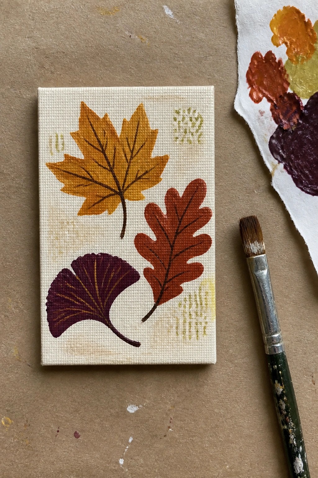 Small canvas with yellow maple, red oak, and purple ginkgo leaves beside paintbrush and palette.