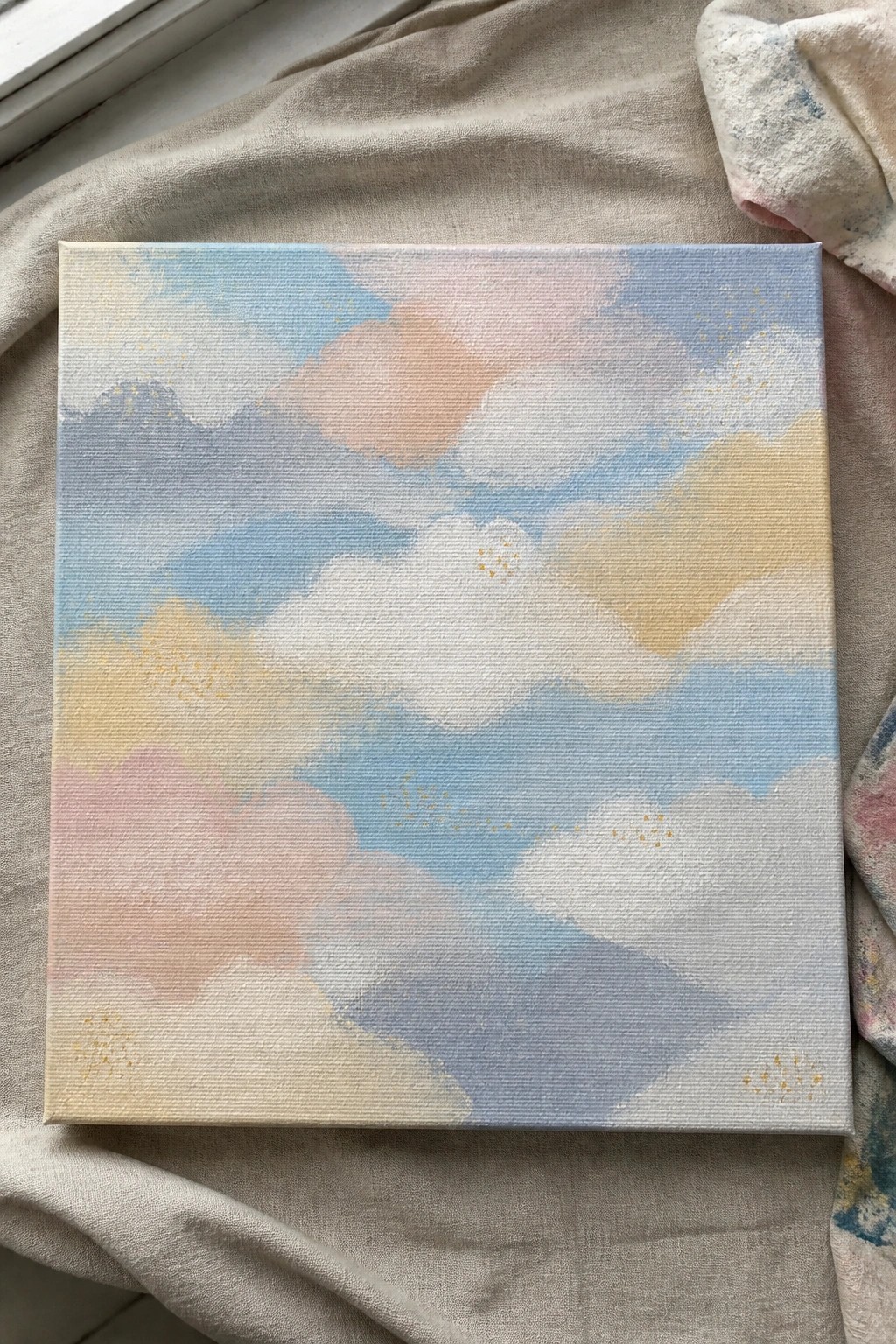 Soft pastel clouds with gold specks on square canvas over beige fabric.