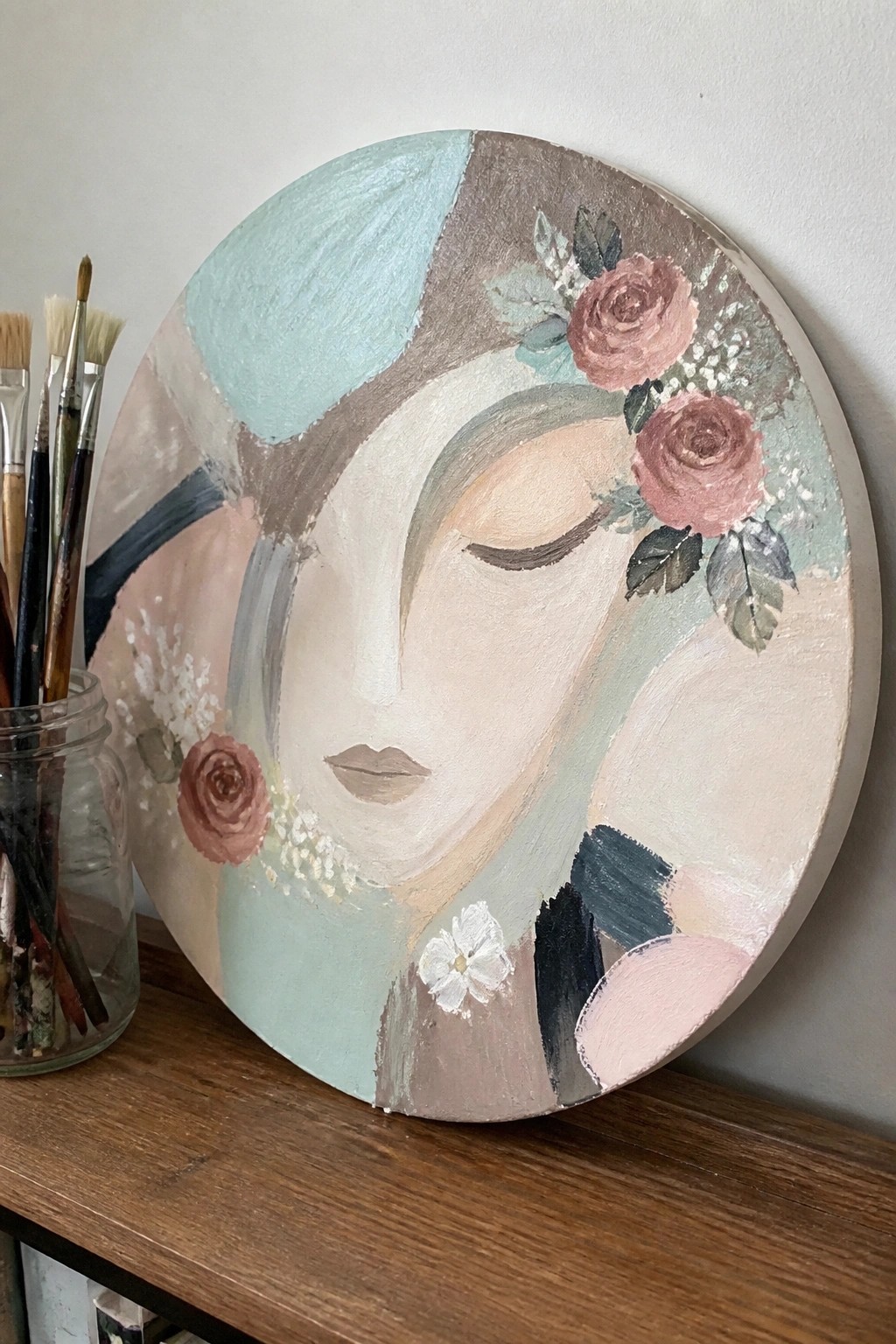 Circular pastel painting of woman's face with pink roses beside paintbrushes on wood