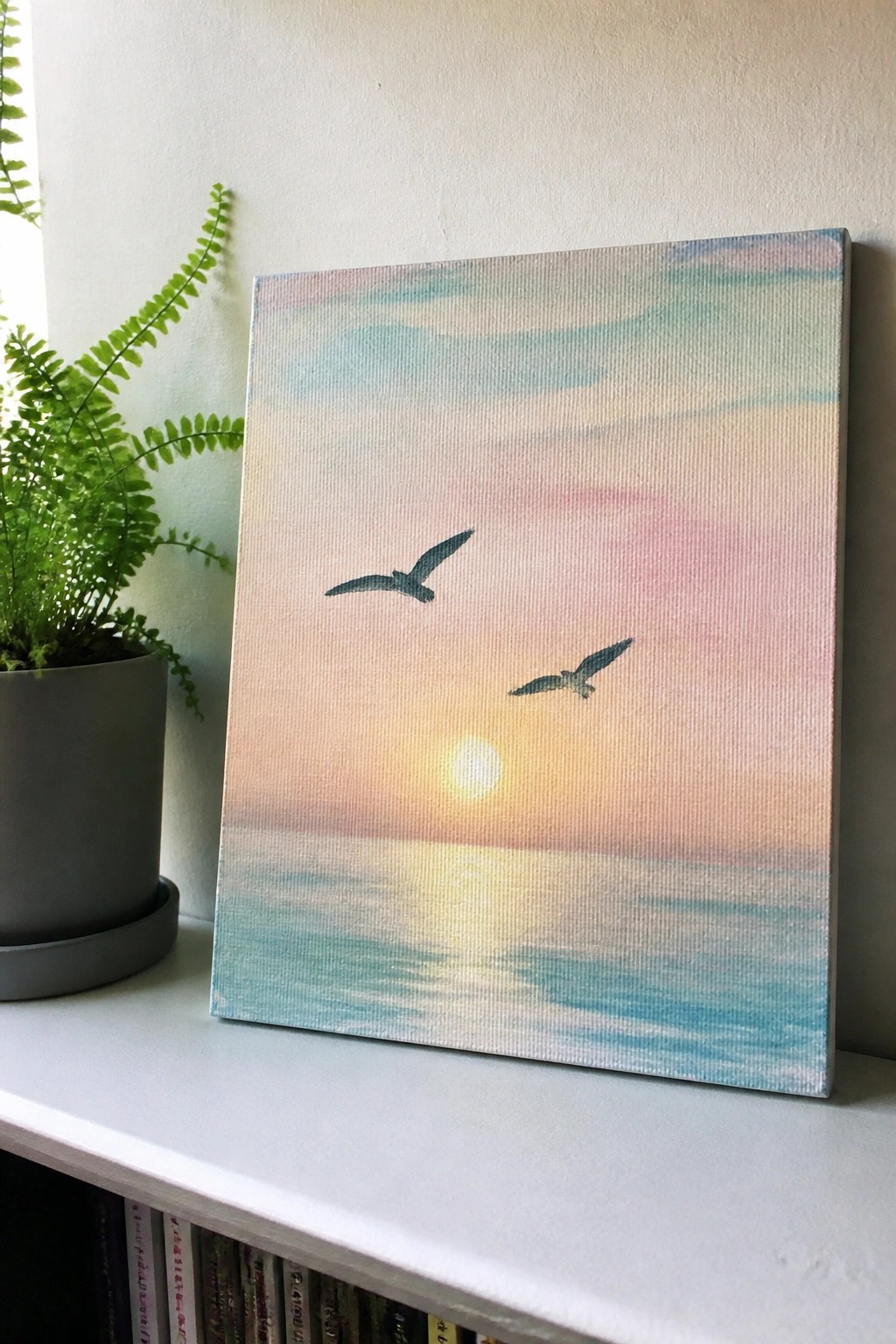 Pastel sunset seascape painting with two birds on white shelf beside fern plant.