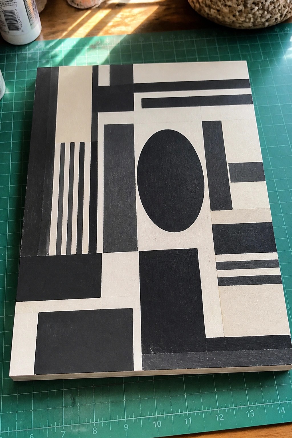 A black and white acrylic painting on a green cutting mat featuring various geometric shapes including rectangles, vertical lines, horizontal lines, and an oval arranged in a grid pattern.