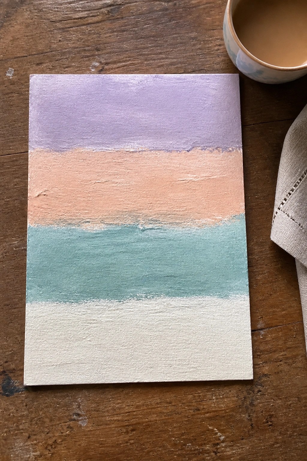 Abstract painting with purple, peach, teal and white stripes beside mug on wood
