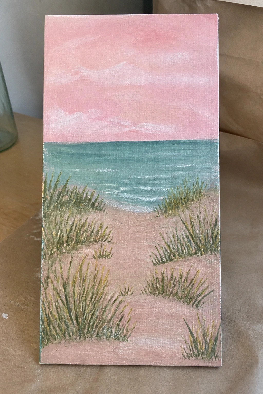 Pink sky over teal ocean with sandy beach and green dune grass painting.