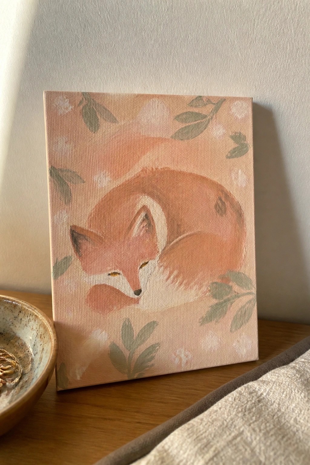 Curled sleeping fox painting in soft orange tones on a wooden table with bowl