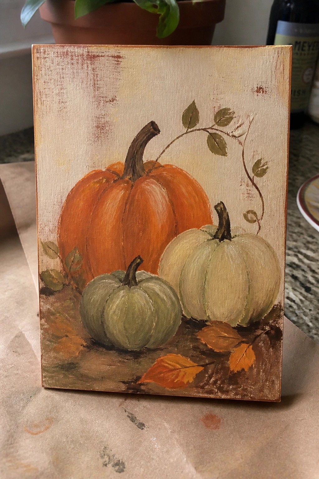 Painting of large orange pumpkin with smaller green and white ones amid fall leaves