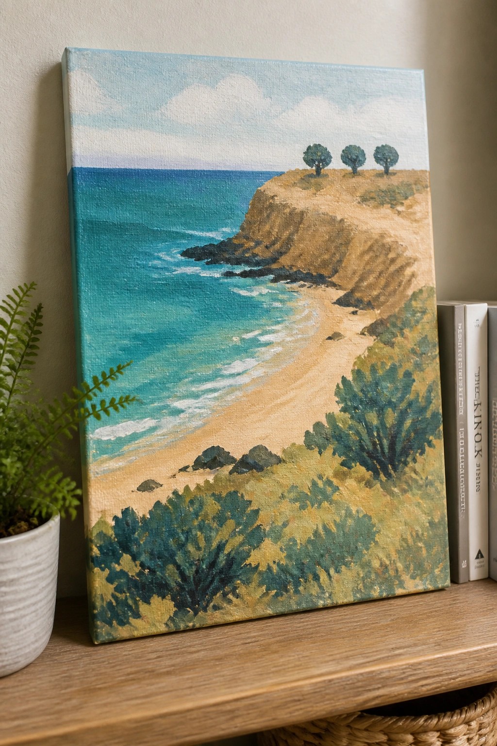 Canvas painting of turquoise ocean, beach and cliffs beside fern and books on shelf