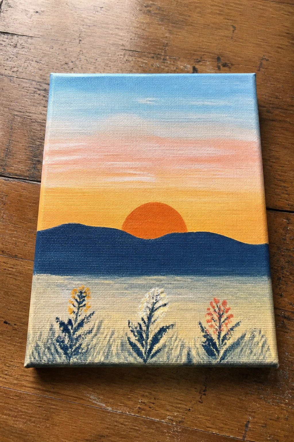 Sunset painting on canvas with orange sun, blue hills, and three wildflowers.