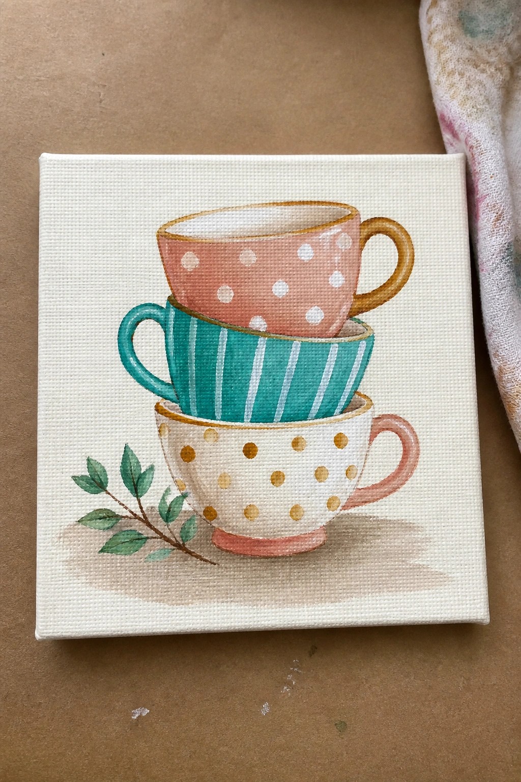 Three stacked teacups with pink polka dots, teal stripes, and white gold dots sit on a white canvas with a small leafy branch and soft shadow.