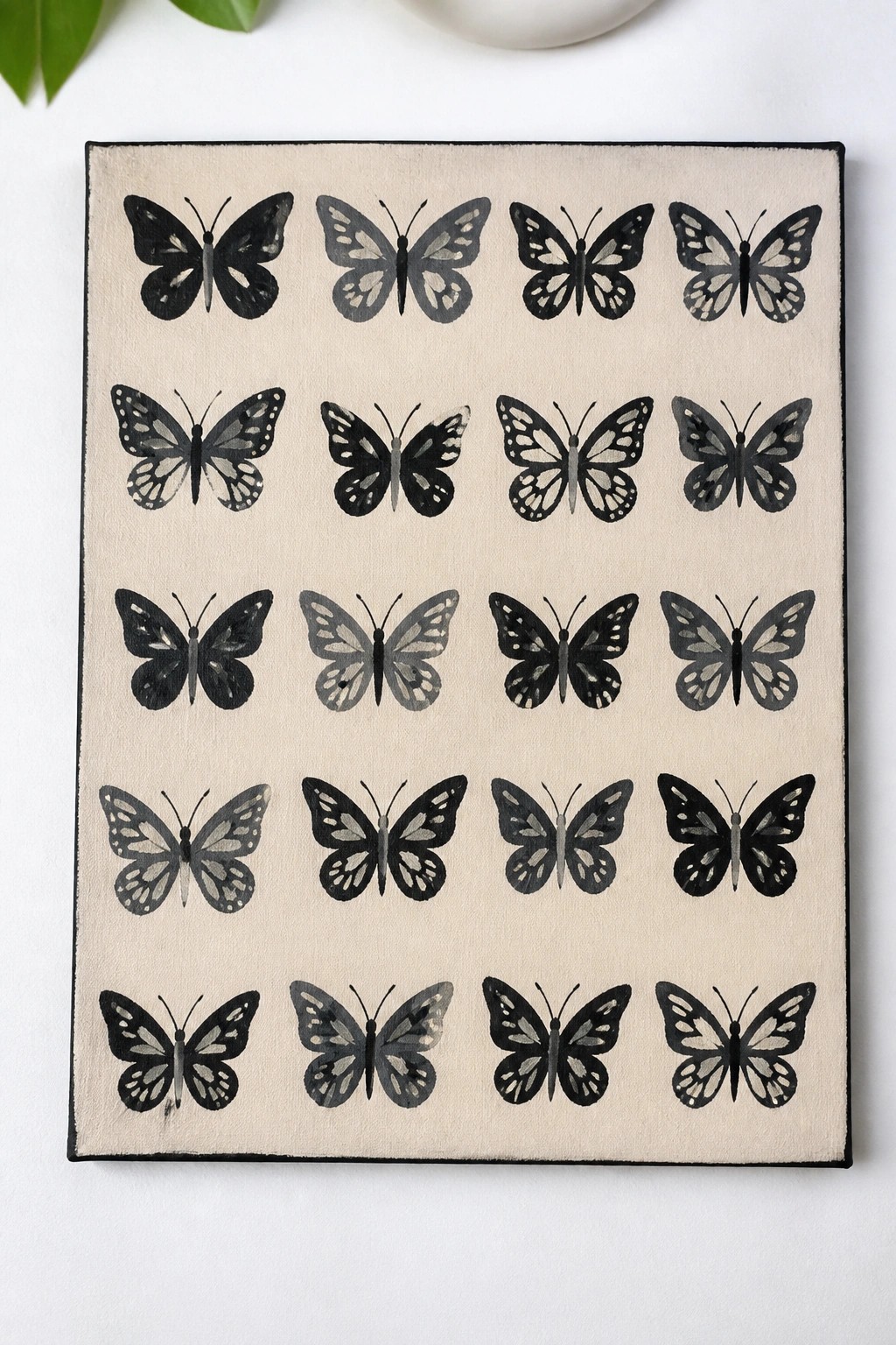 Rectangular canvas with rows of black and gray butterfly paintings on beige background