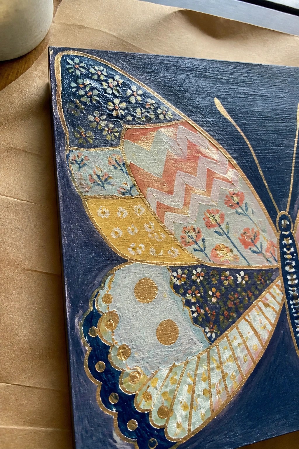 Ornate butterfly wing painting with floral patterns, chevrons, polka dots, and gold accents on blue.