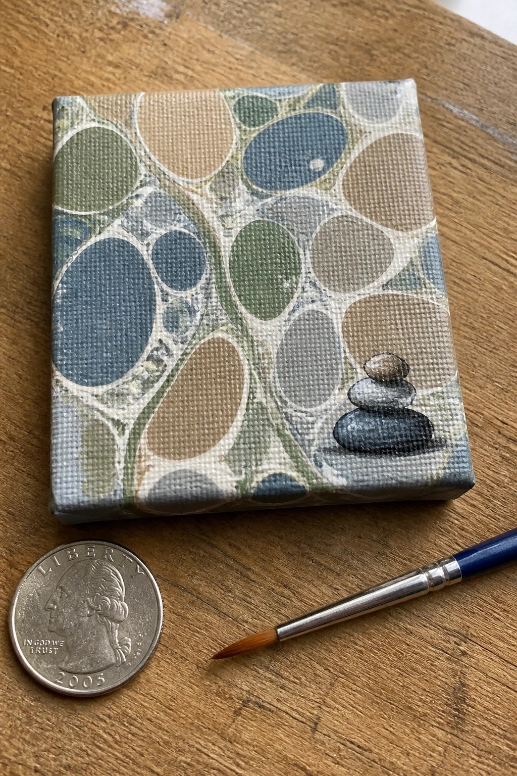 Small canvas with circle pattern and stacked stones beside quarter and paintbrush on wood