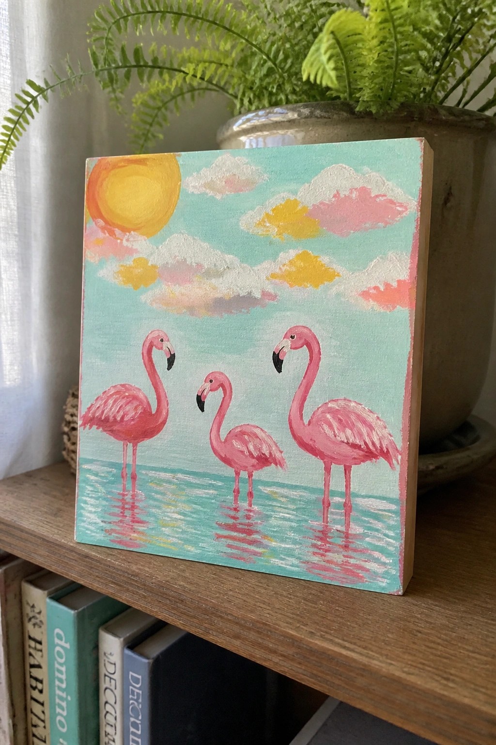 Three pink flamingos wading in turquoise water beneath a yellow sun and clouds