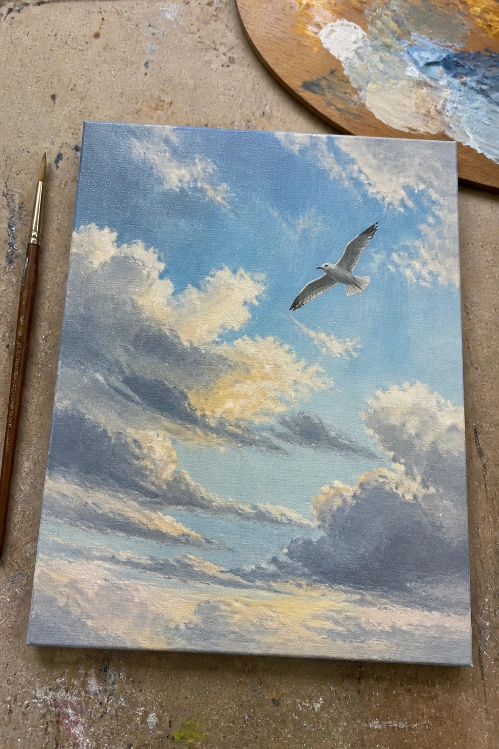 Painting of seagull soaring over cloudy blue ocean, beside paintbrush and palette.