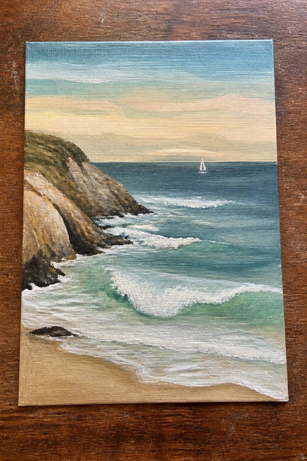 Painting of rocky cliffs, sandy beach, turquoise waves, and distant sailboat at sunset.