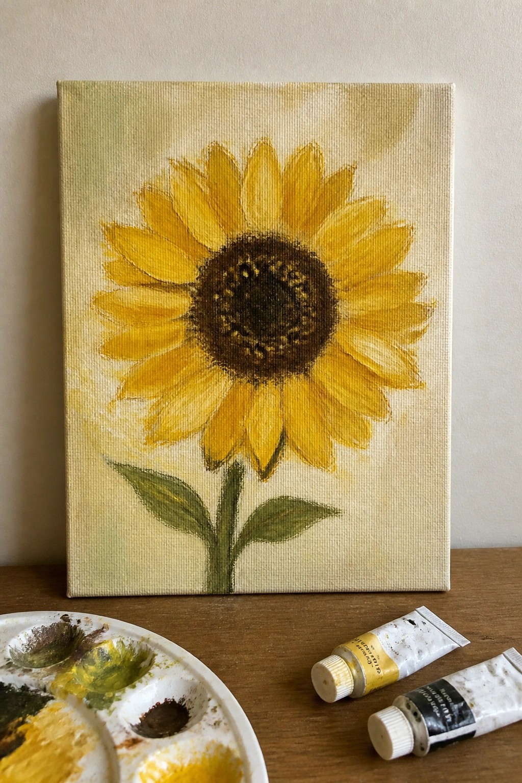 Sunflower painting on canvas beside paint palette and tubes on wooden table
