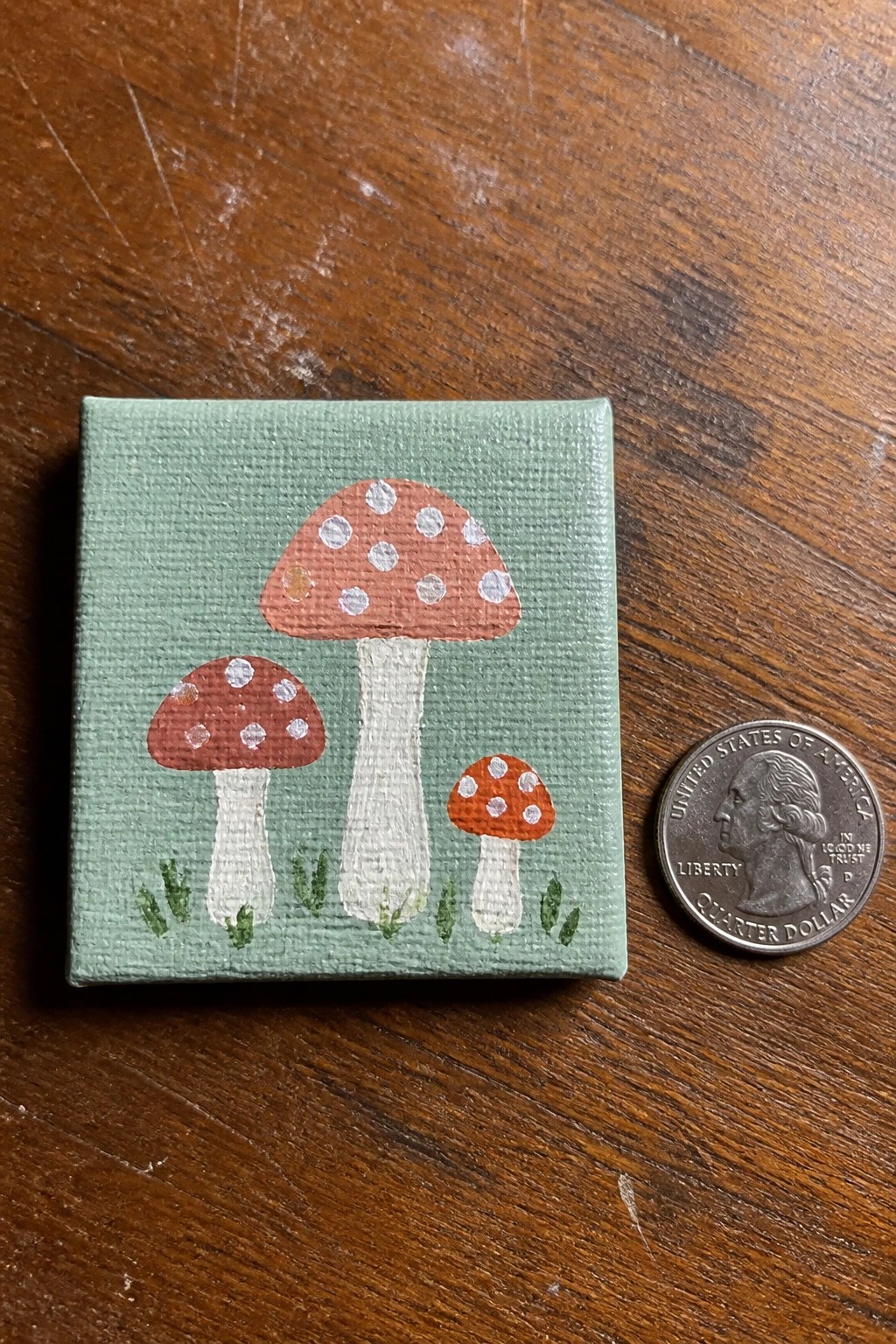 A small square canvas with three mushrooms of different sizes painted on a light green background sits next to a US quarter on a wooden table.