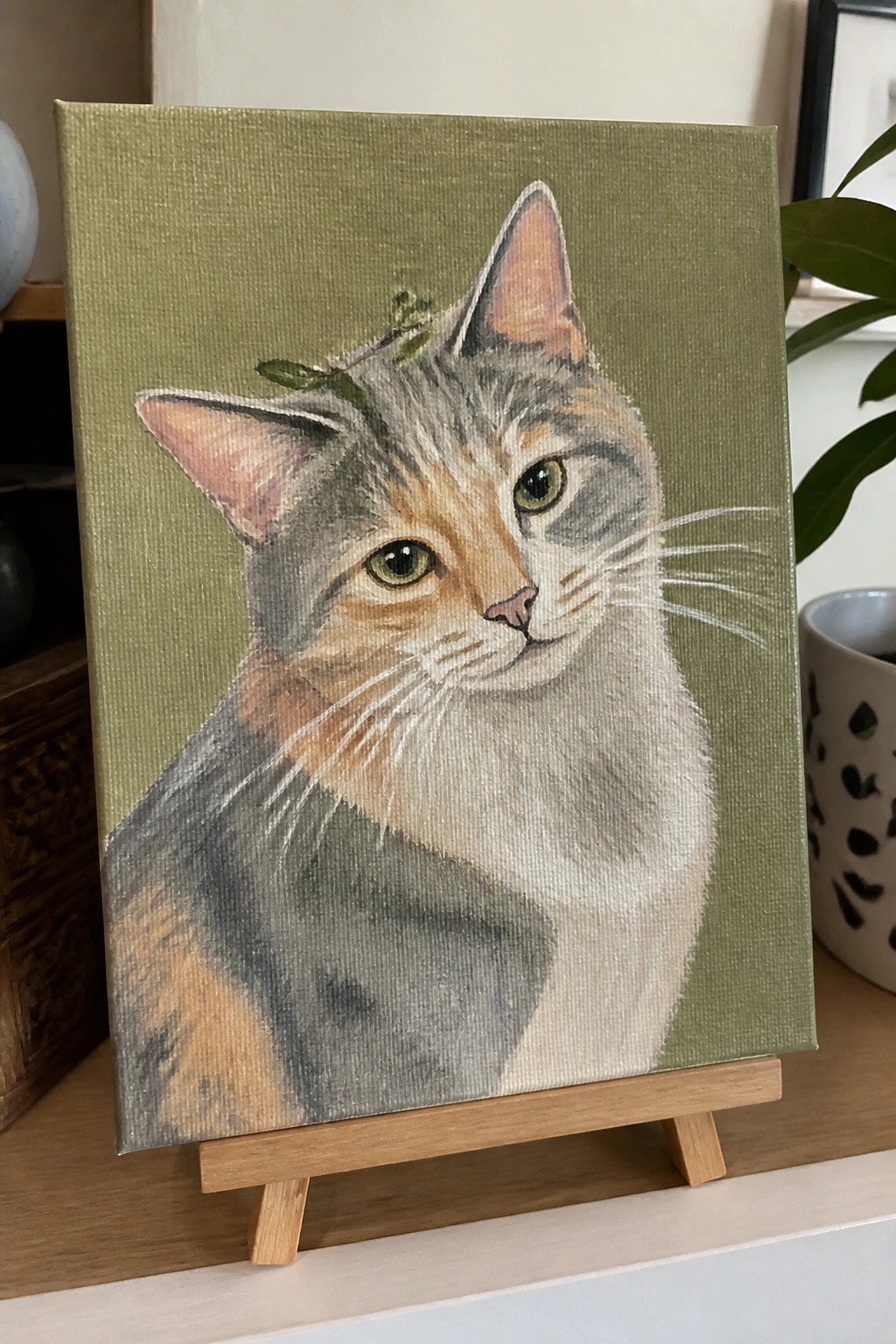 Calico cat with green eyes on sage green canvas atop wooden easel