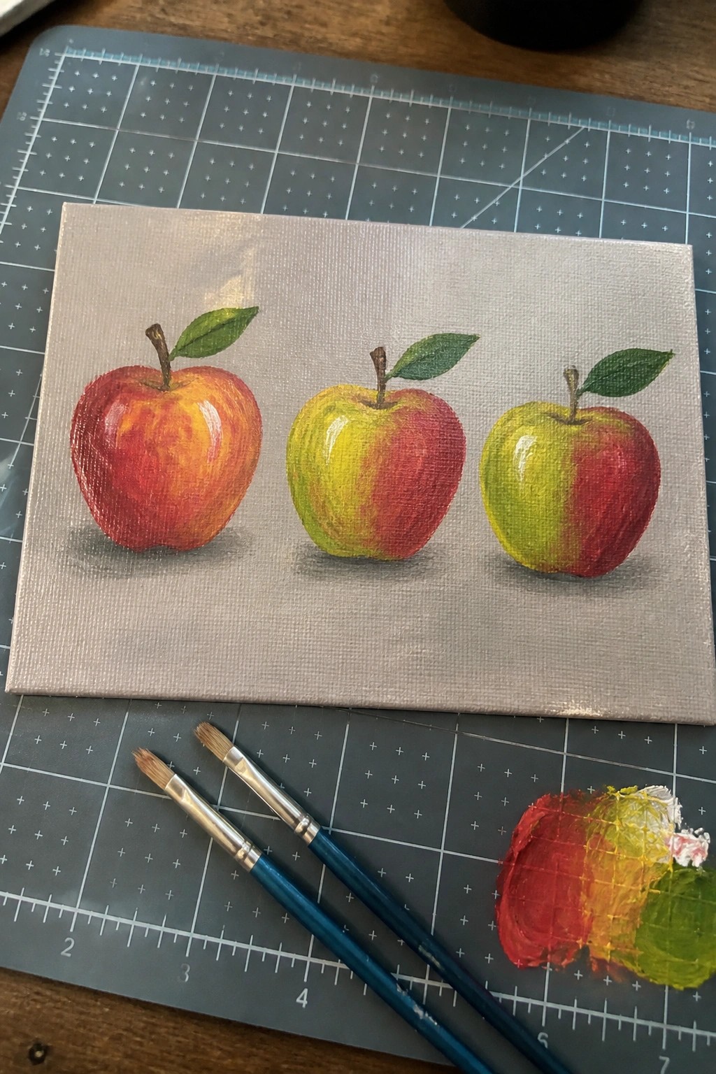 Three painted apples in red, yellow, and green on canvas with brushes and palette