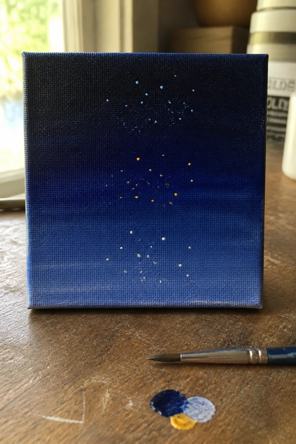 Small blue canvas with white and gold stars, paintbrush and paints on wood table
