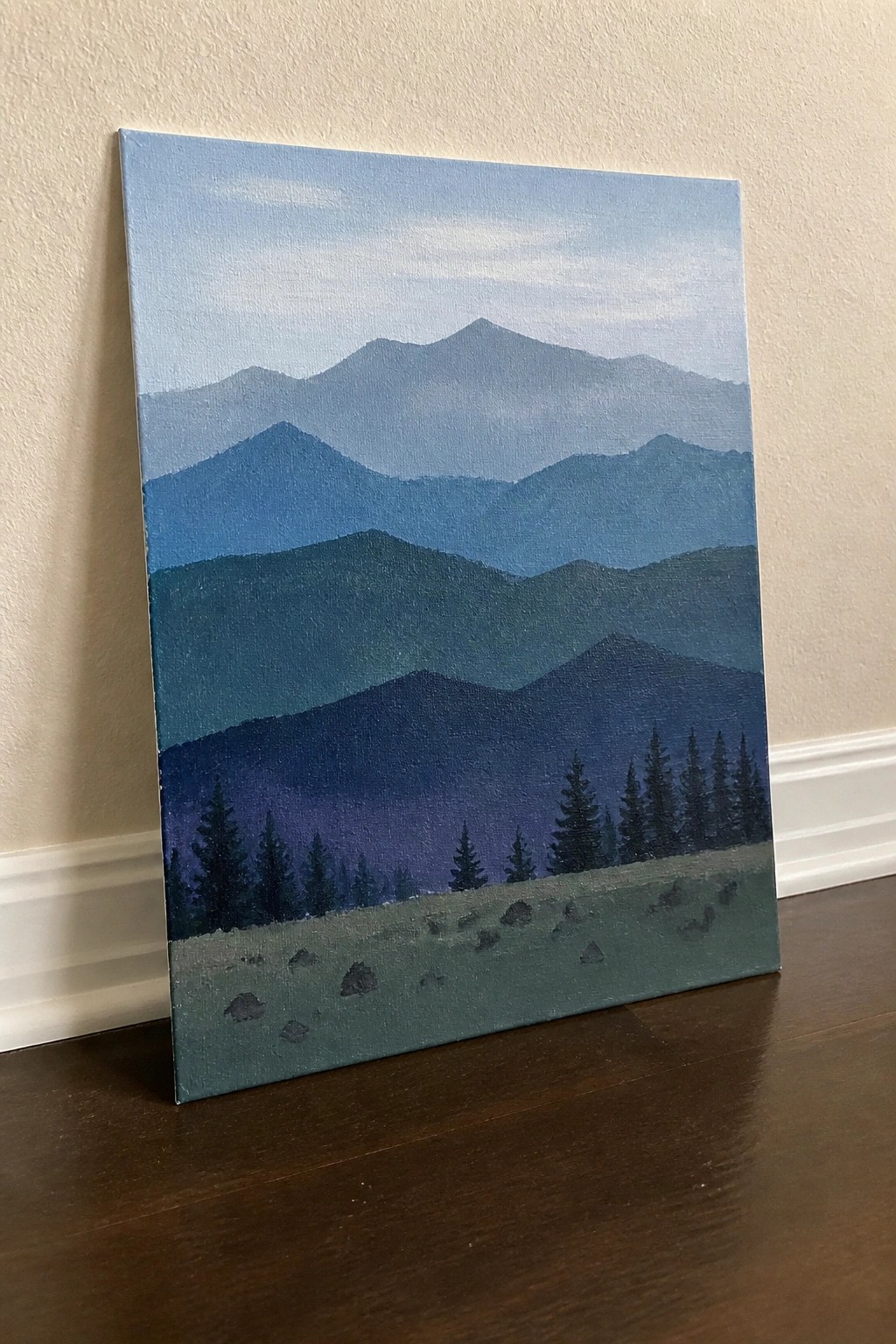 Layered blue mountains with silhouetted pine trees over rocky green meadow on canvas.