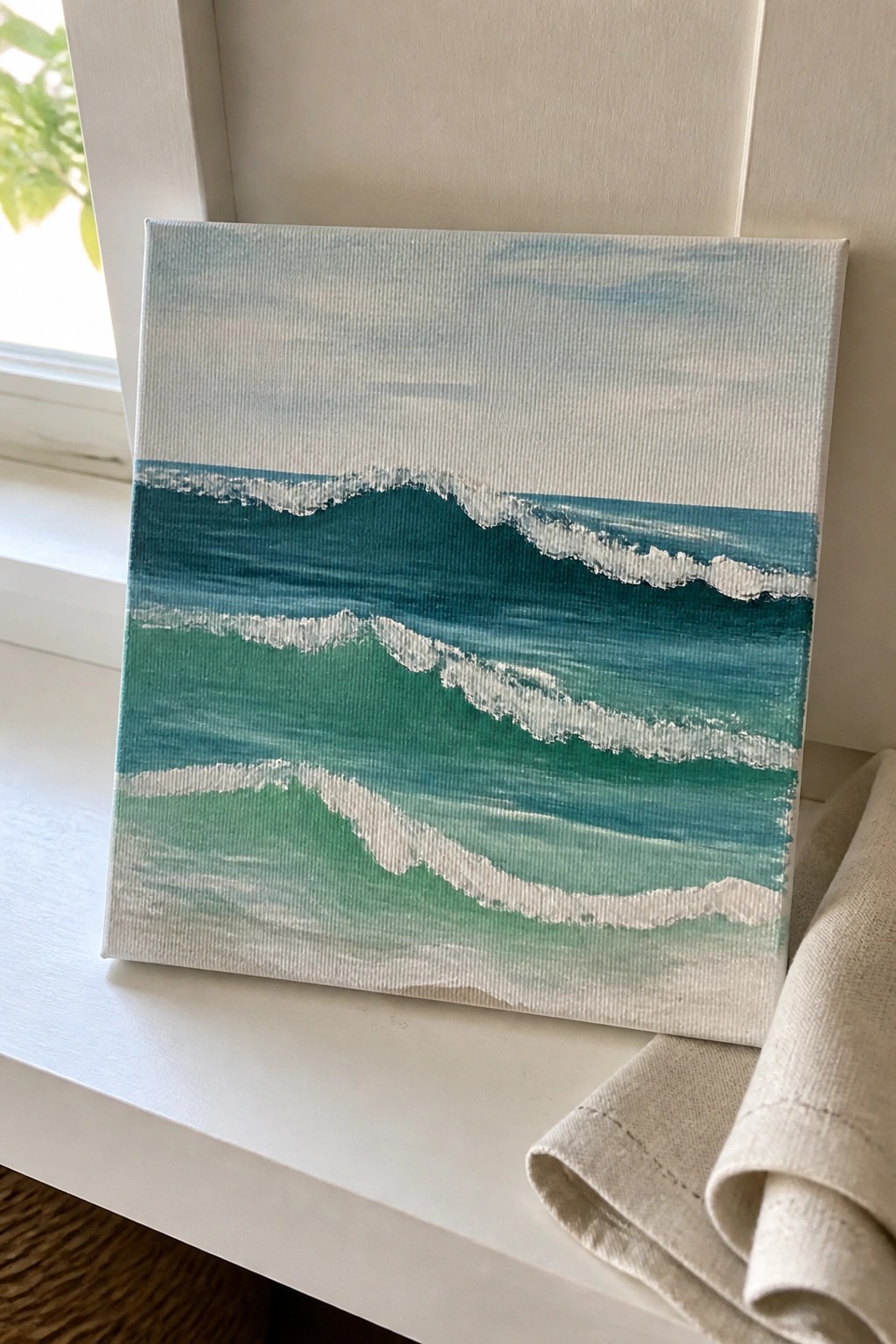 Blue-green wave painting on canvas beside white windowsill with beige cloth