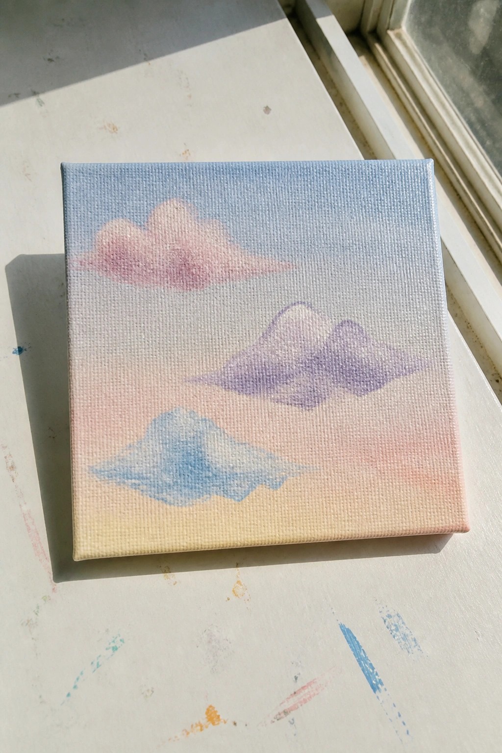 Small square canvas with pastel cloud painting on paint-splattered windowsill in sunlight.
