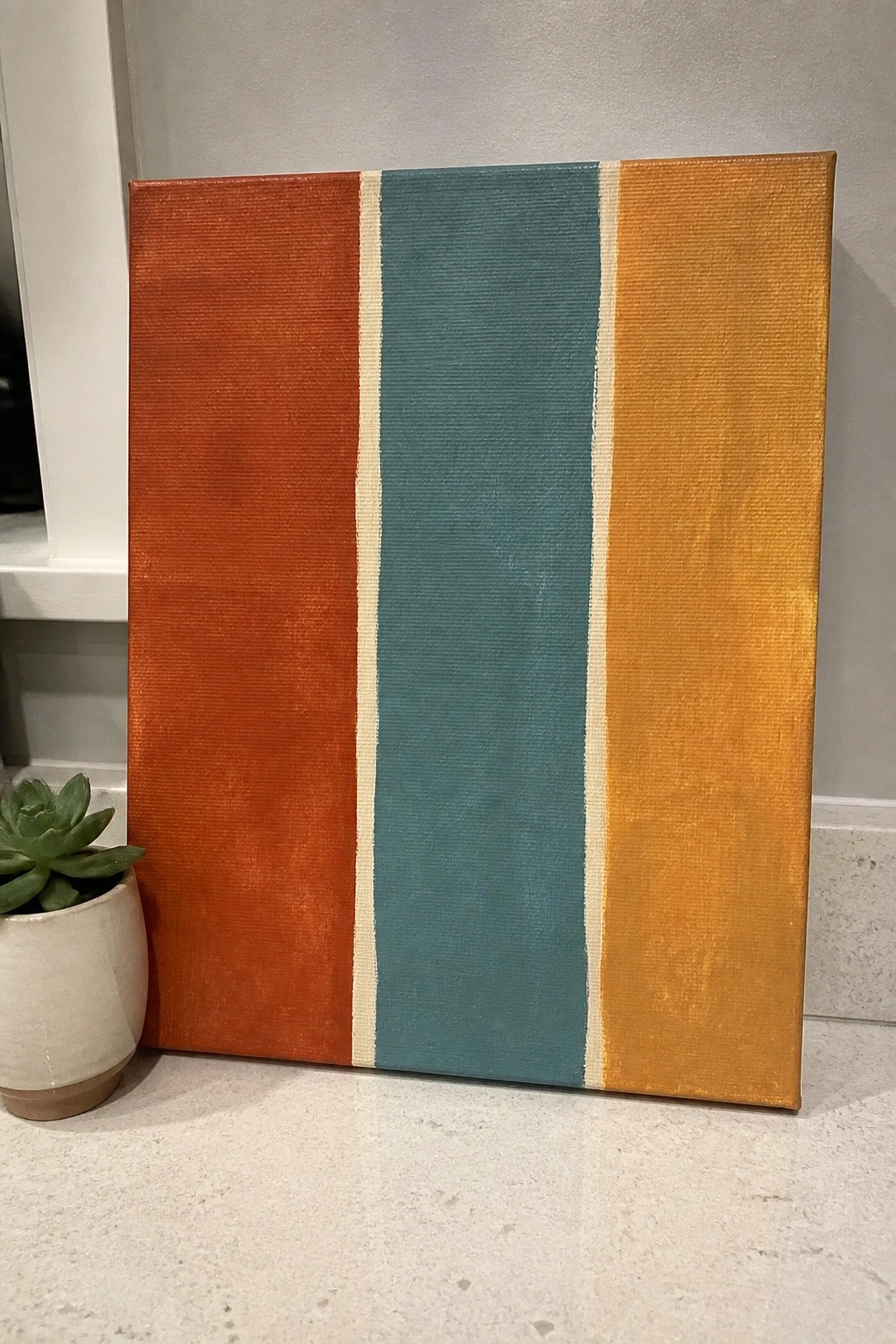 Striped canvas painting in orange, teal, and yellow beside potted succulent on counter