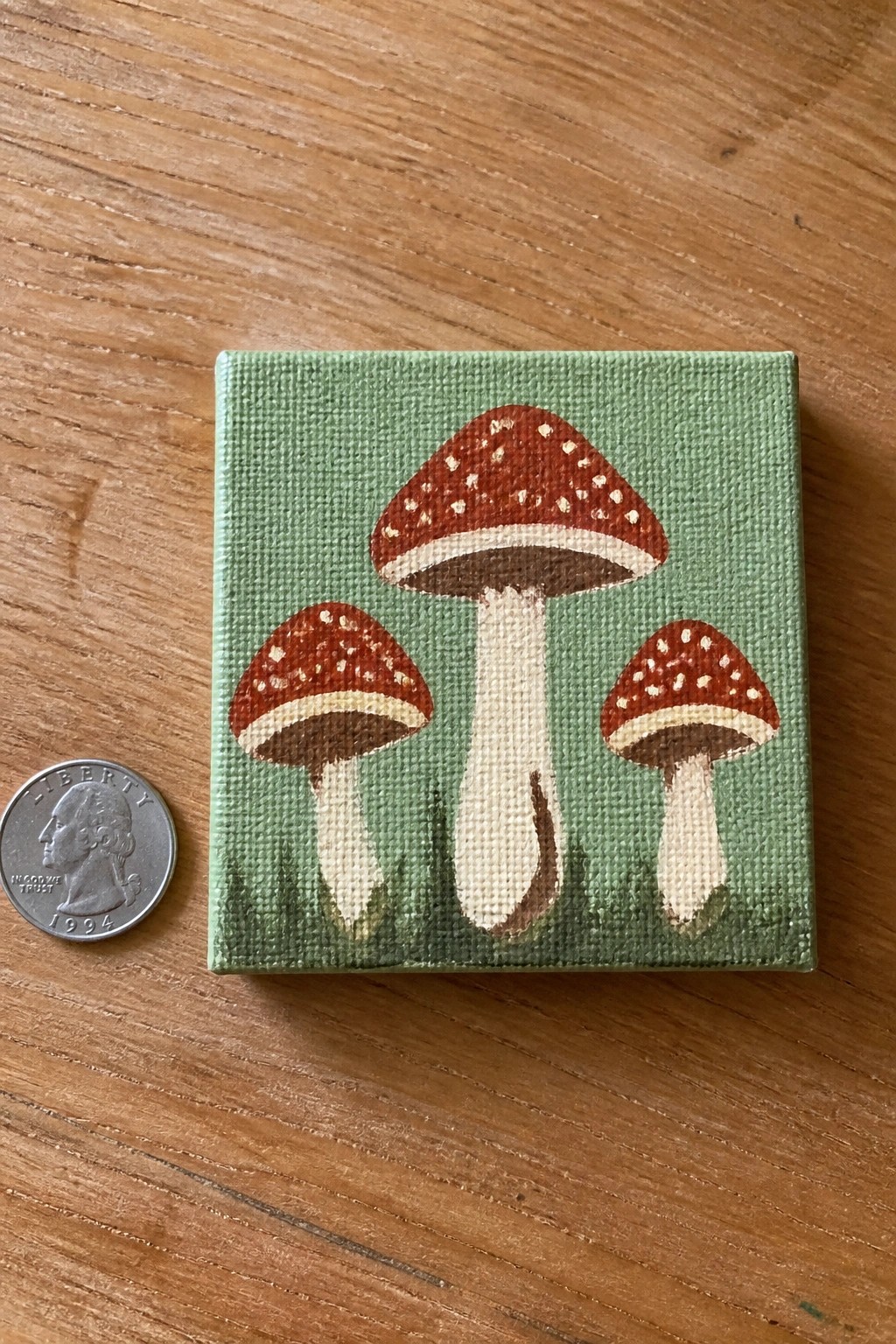 Miniature canvas painting of three red spotted mushrooms beside a US quarter on wood.