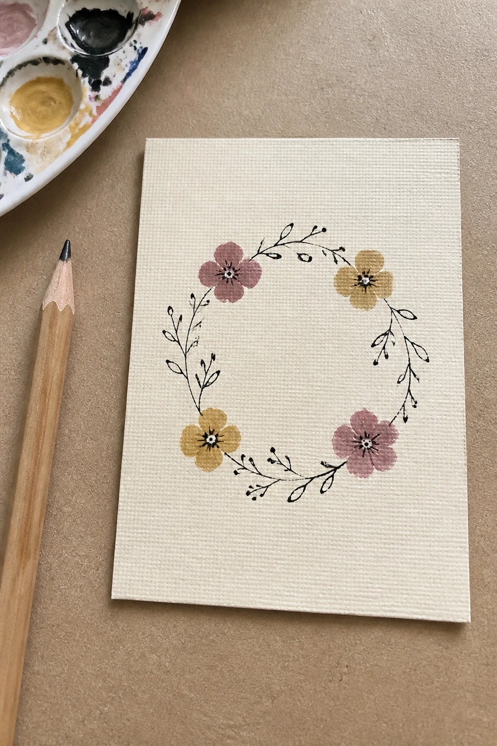 A small white canvas shows a circular wreath of four flowers in yellow and muted pink connected by black line branches with tiny leaves.