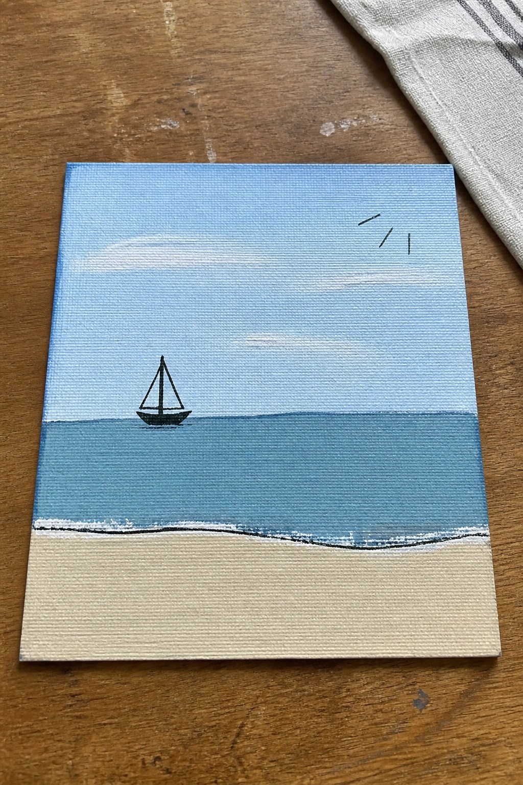 Small canvas painting of sailboat on blue sea above sandy beach with sun on wood table