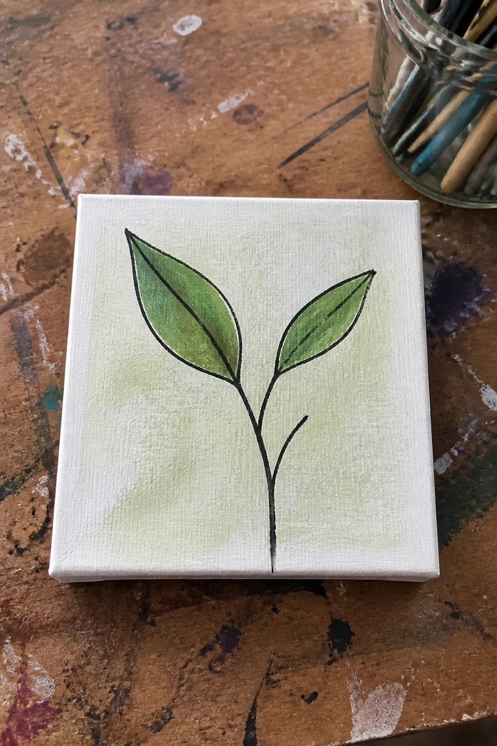 A small white canvas shows a black-outlined stem with two green leaves on a light green wash background resting on a wooden table beside a jar of brushes.