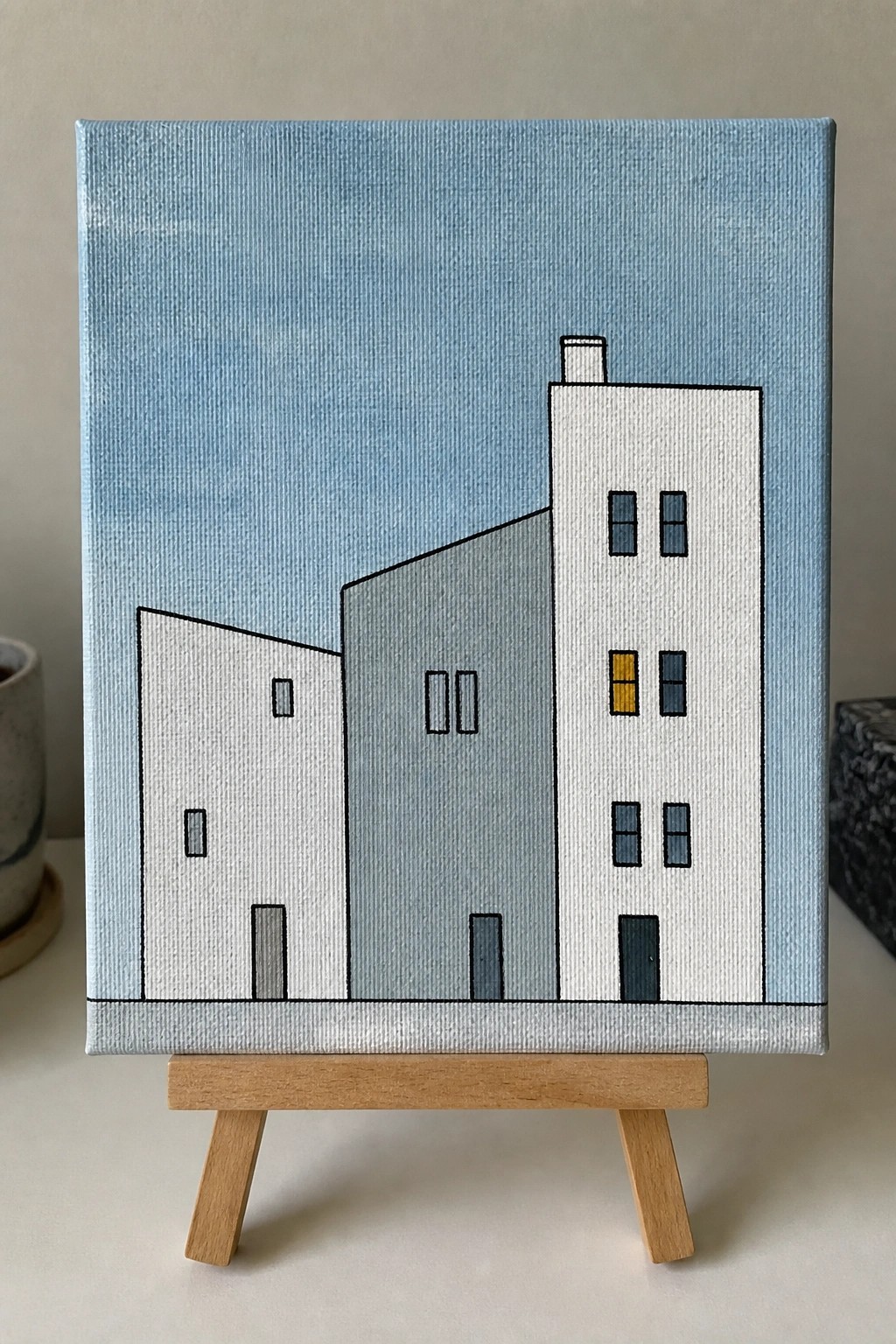 Minimalist painting of three white and gray buildings on wooden easel.