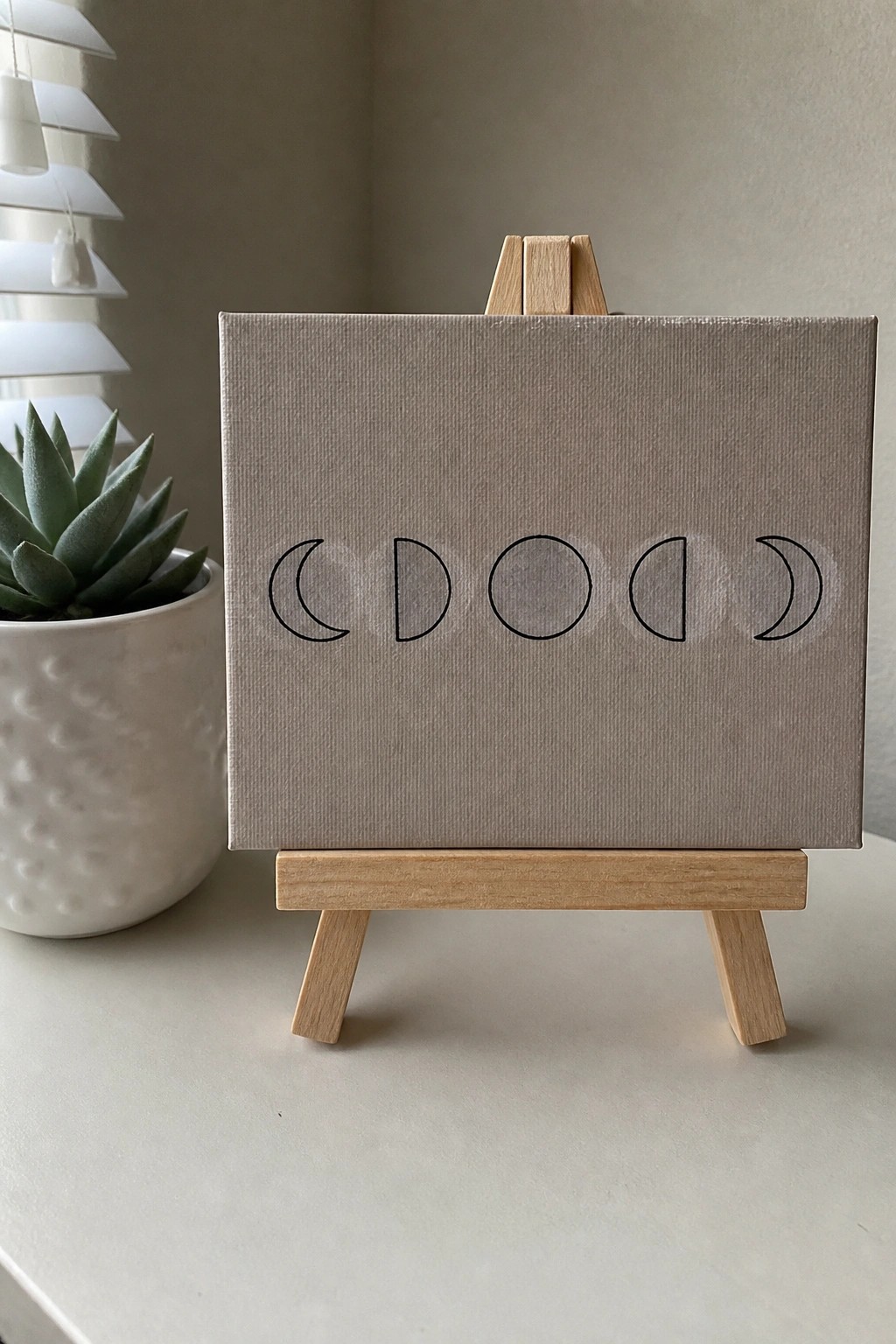 A small beige canvas on a wooden easel shows five black line drawings of moon phases arranged in a row.