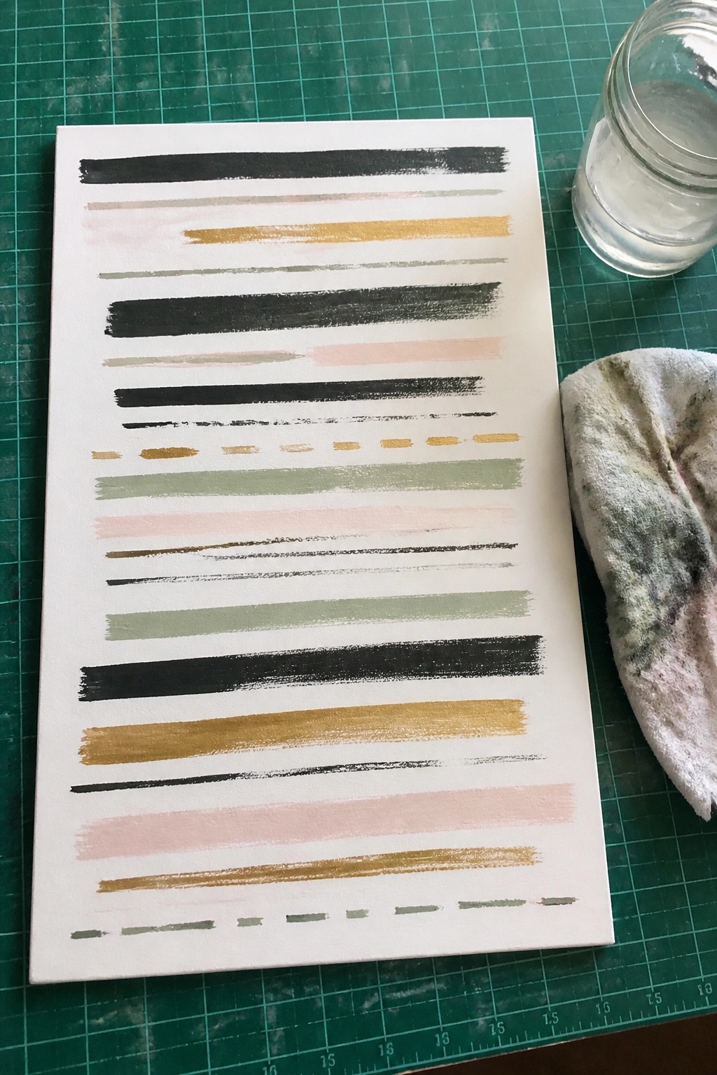 A white canvas on a green cutting mat shows multiple horizontal brush strokes in black, gold, pink, green, and beige acrylic paint next to a jar of water and a rag.