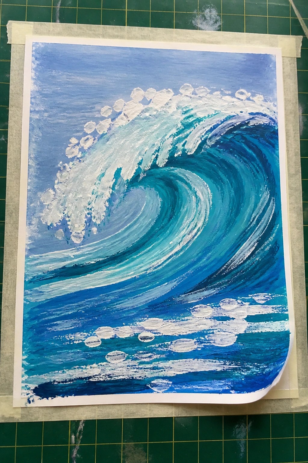Acrylic painting of a large curling blue ocean wave with white foam on top and smaller foam lines in the water below, taped to a green cutting mat.