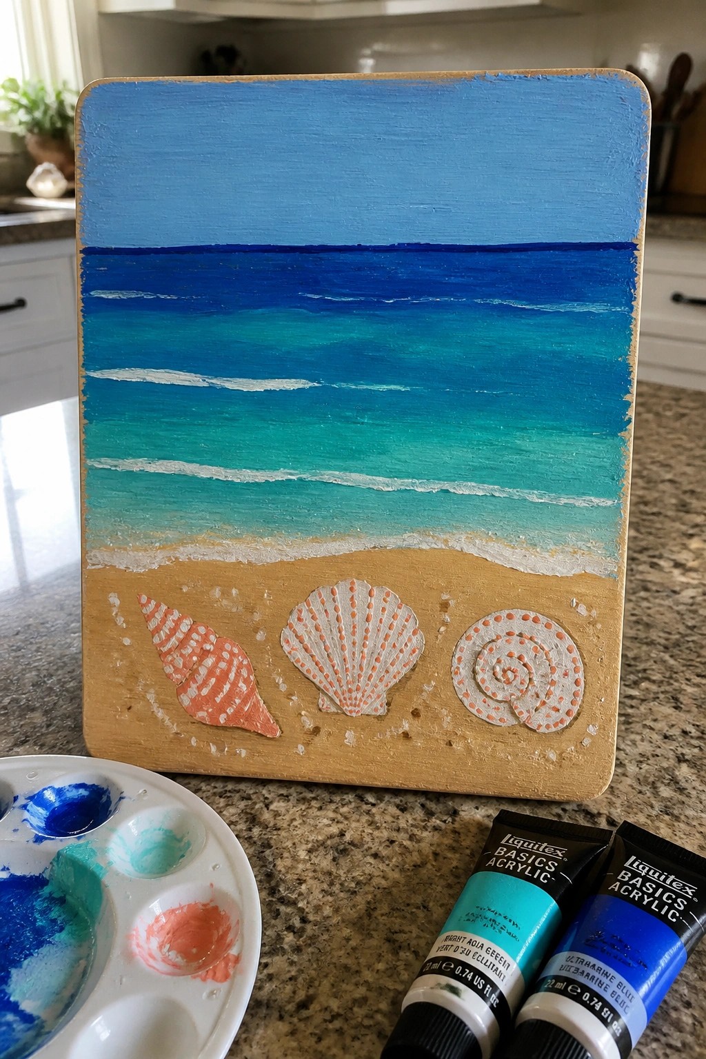 Wood panel beach scene with ocean waves, sand, seashells, paint palette and tubes on counter.