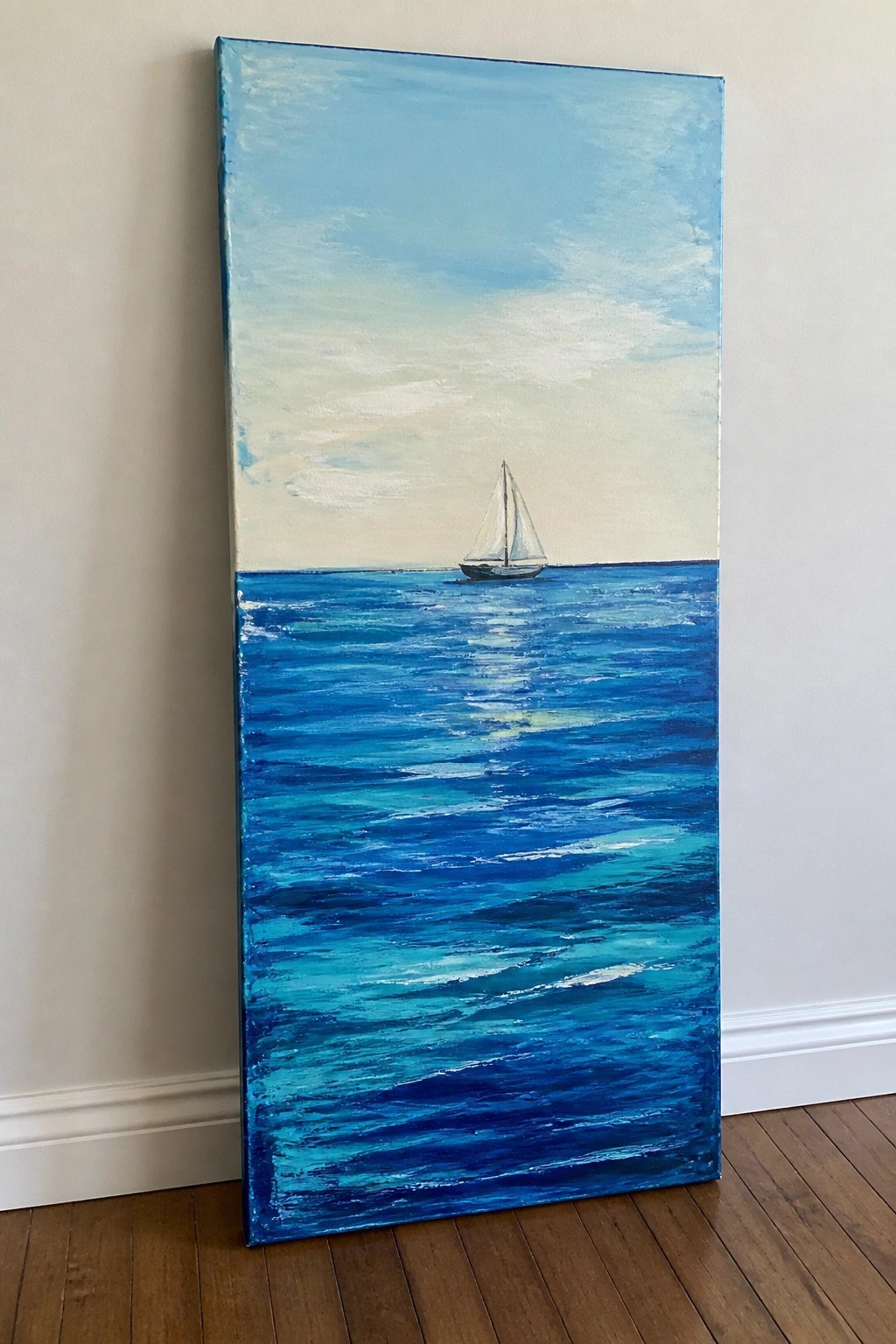 Vertical blue seascape painting of sailboat on ocean, leaning against wall on wood floor