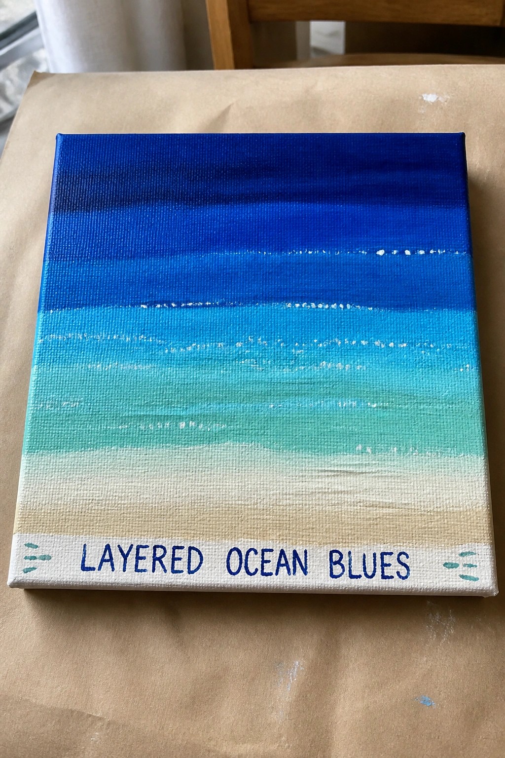 Acrylic canvas with horizontal layers of blue paint fading from dark navy at the top through turquoise to beige at the bottom, with the words Layered Ocean Blues painted along the lower edge.
