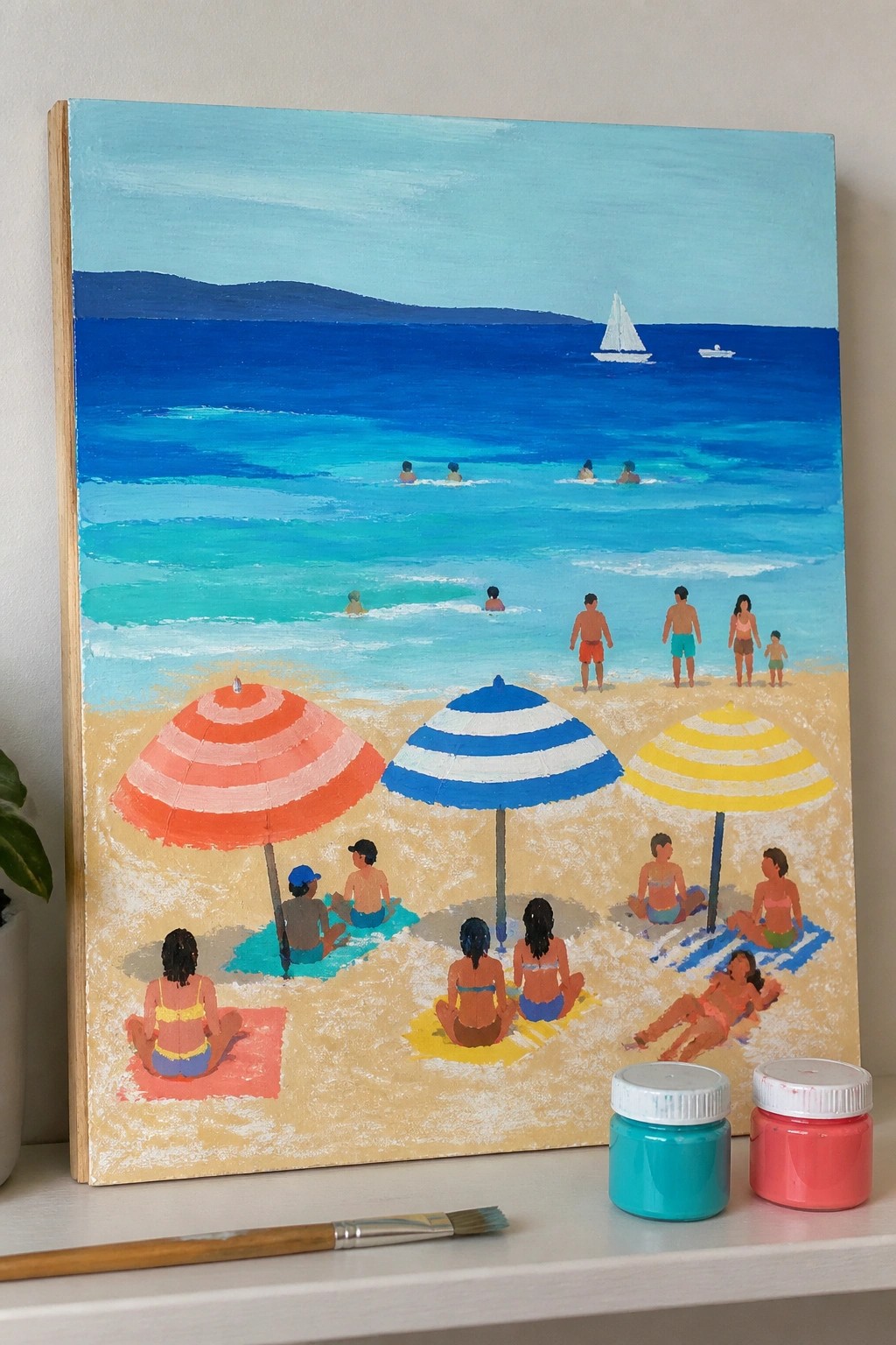 An acrylic painting of a beach with layered blue ocean, yellow sand, three striped umbrellas, people on towels, swimmers, sailboats, and paint jars plus a brush visible below the canvas.