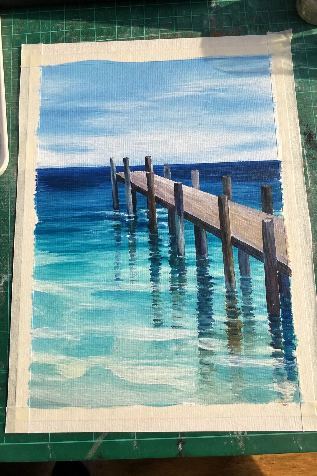 Acrylic painting of a wooden pier extending into blue ocean water with turquoise shallows and a light blue sky, on paper taped to a green mat.
