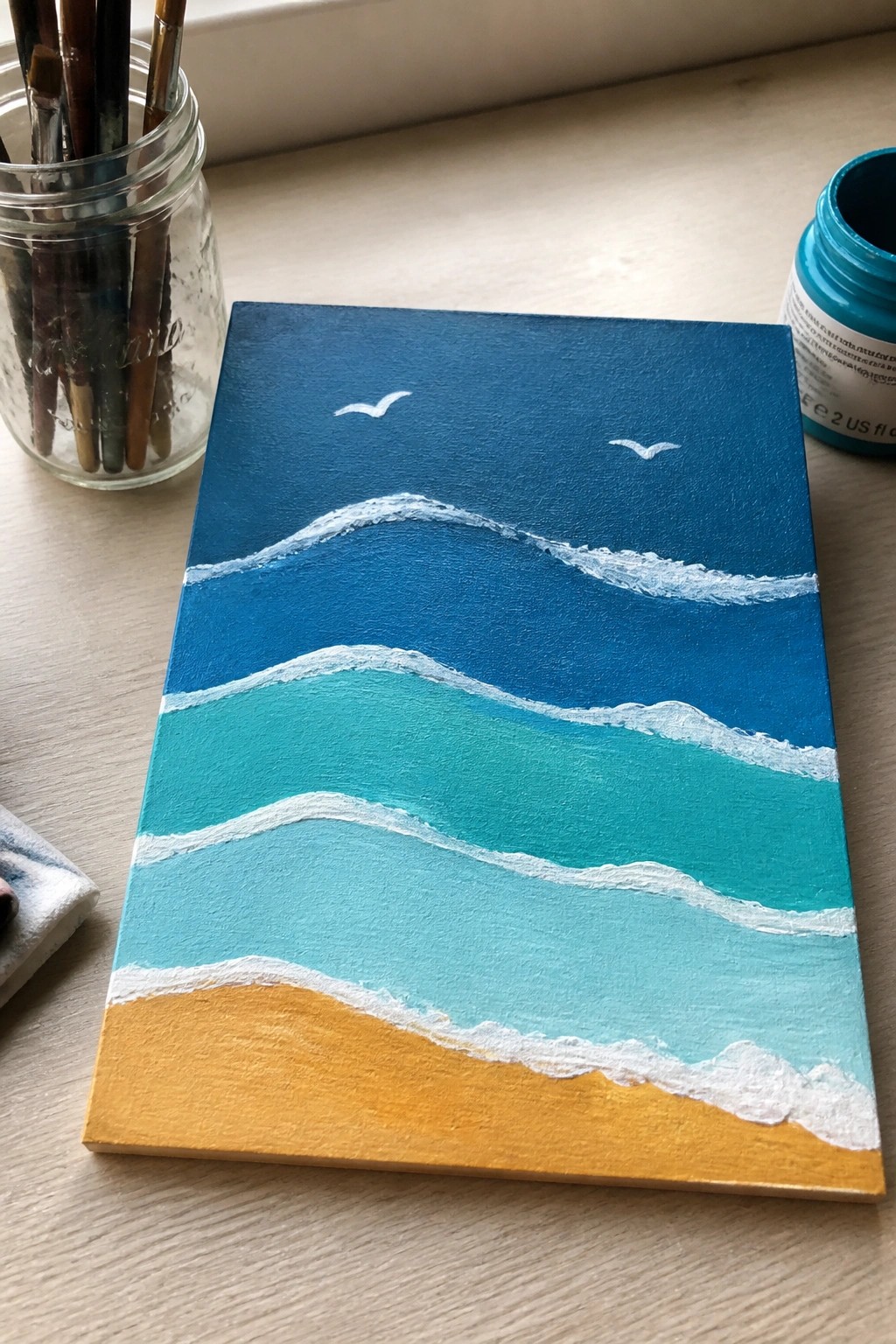 Seascape painting with blue waves, white foam, yellow sand, beside brushes and paint jar.