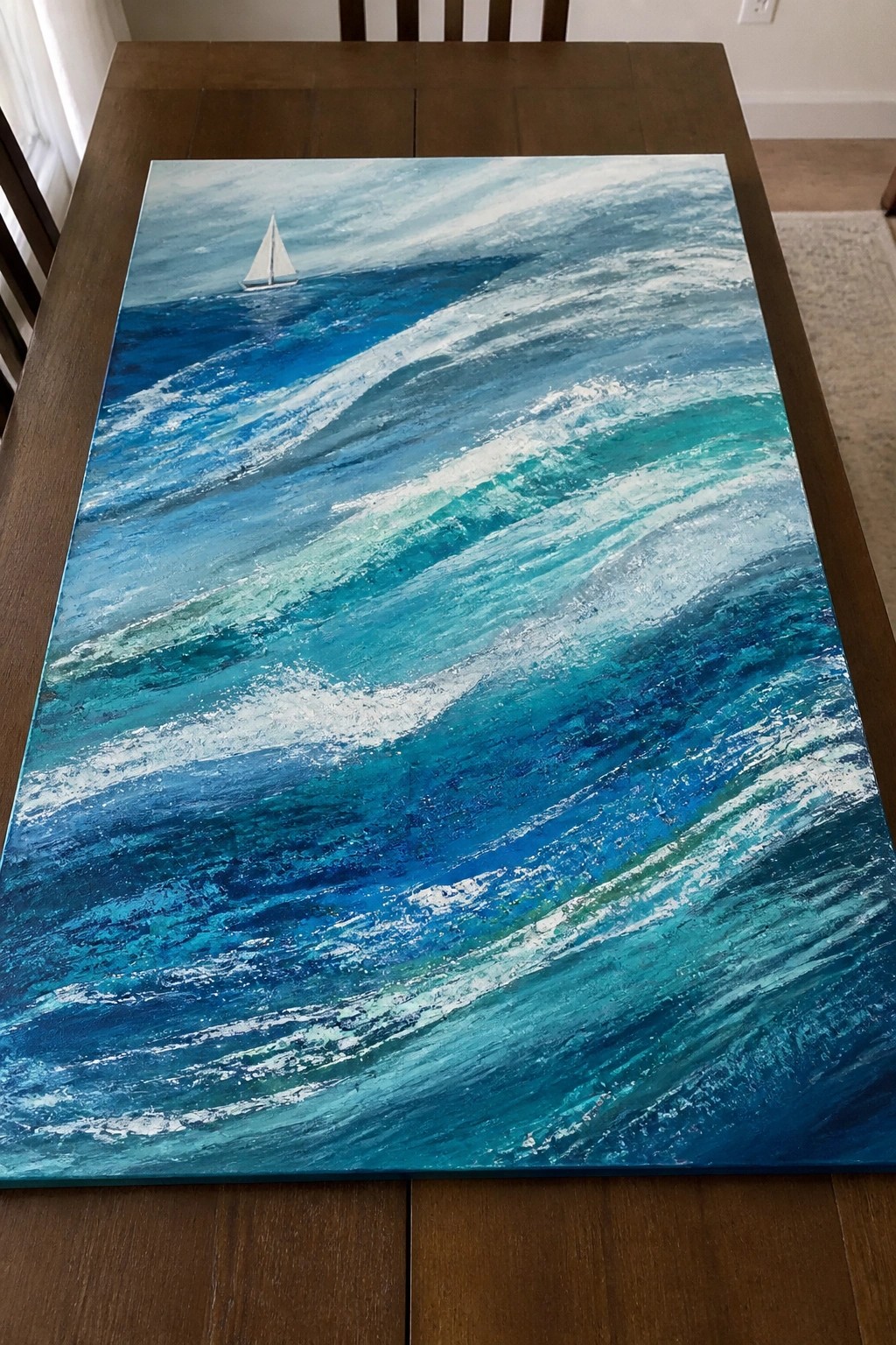 Textured painting of blue-green ocean waves with small sailboat on wooden table