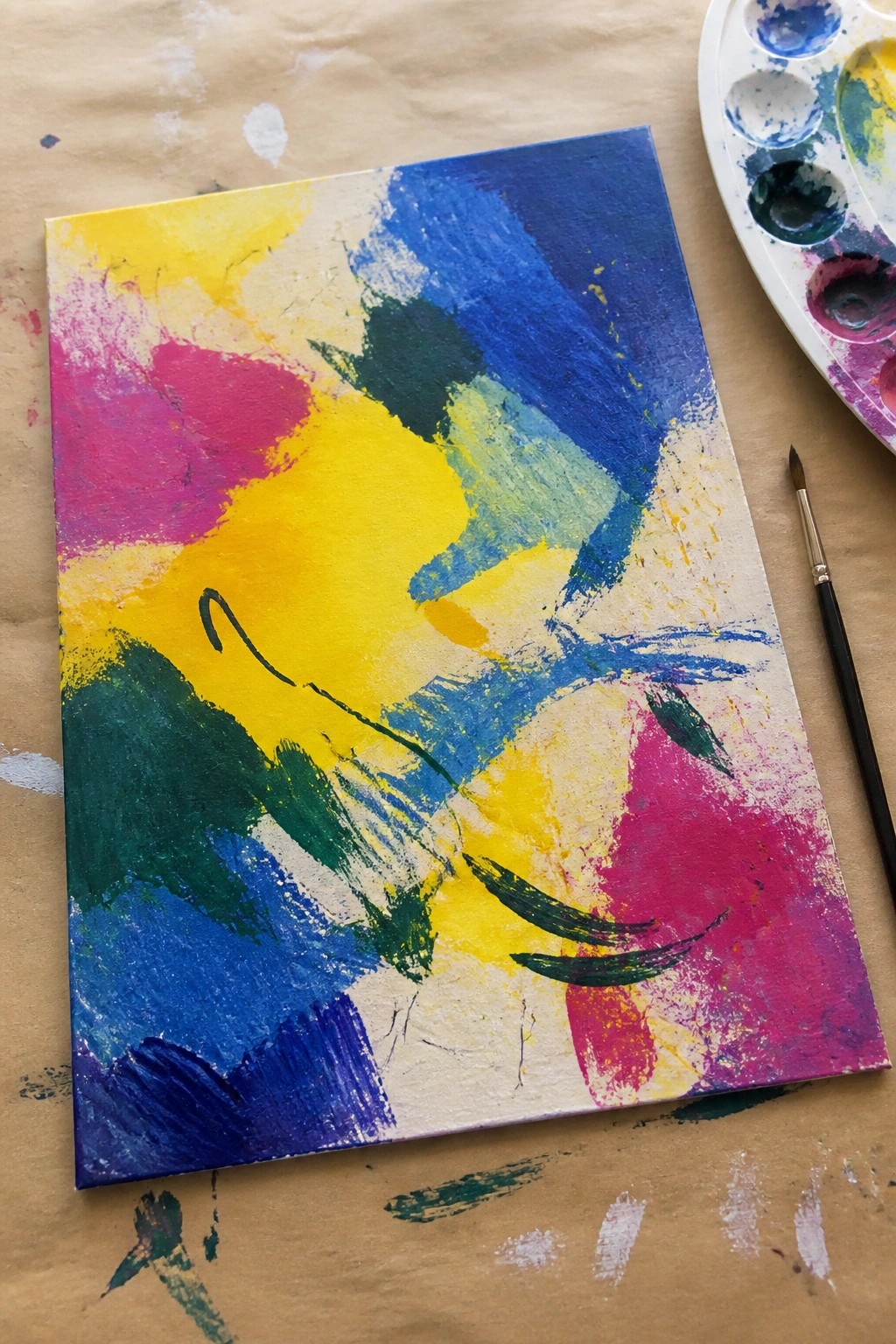 Abstract canvas with bold blue, yellow, pink, and green brushstrokes beside palette and brush.