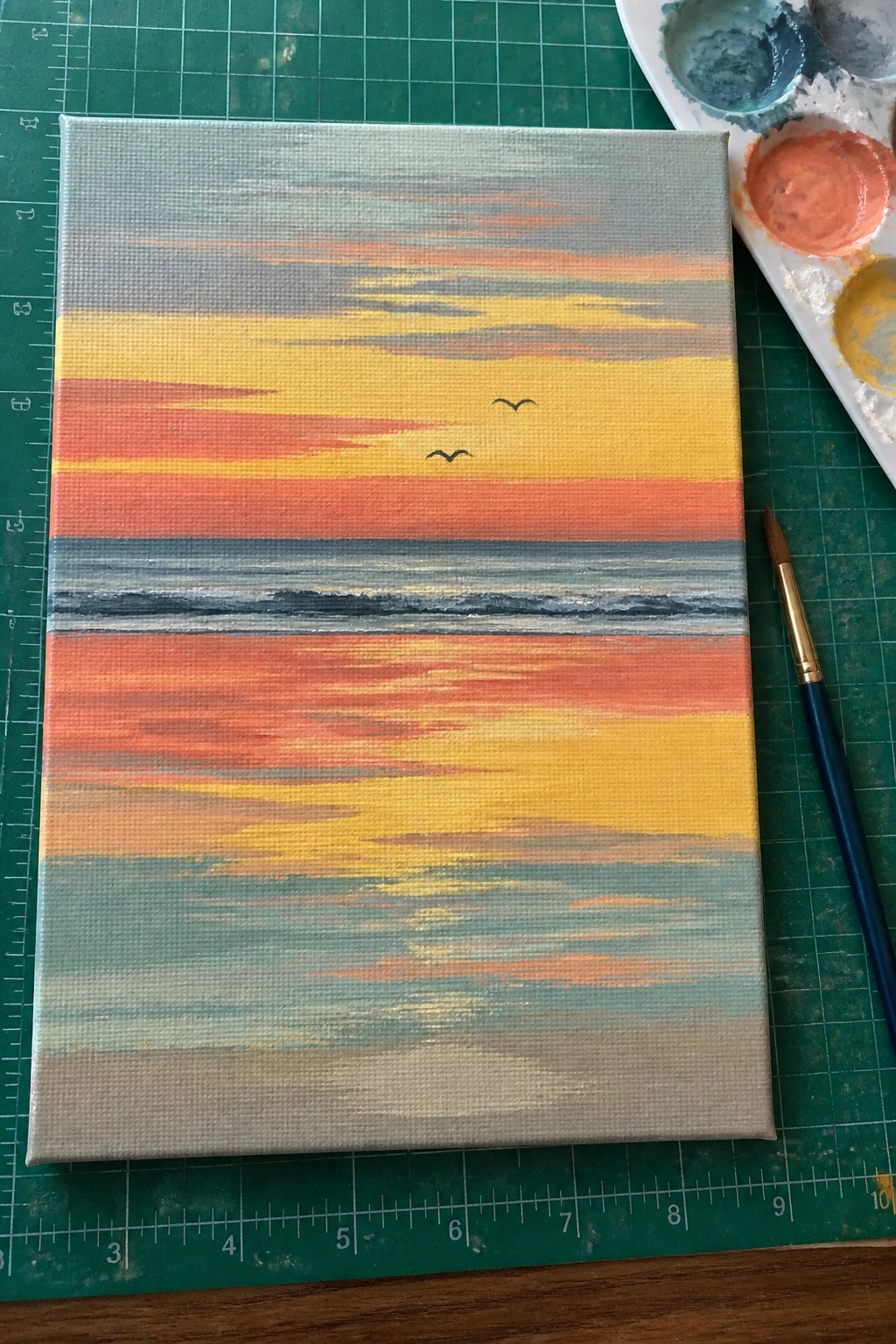 Abstract sunset painting on canvas with orange-yellow stripes, two birds, beside palette and brush on green mat.