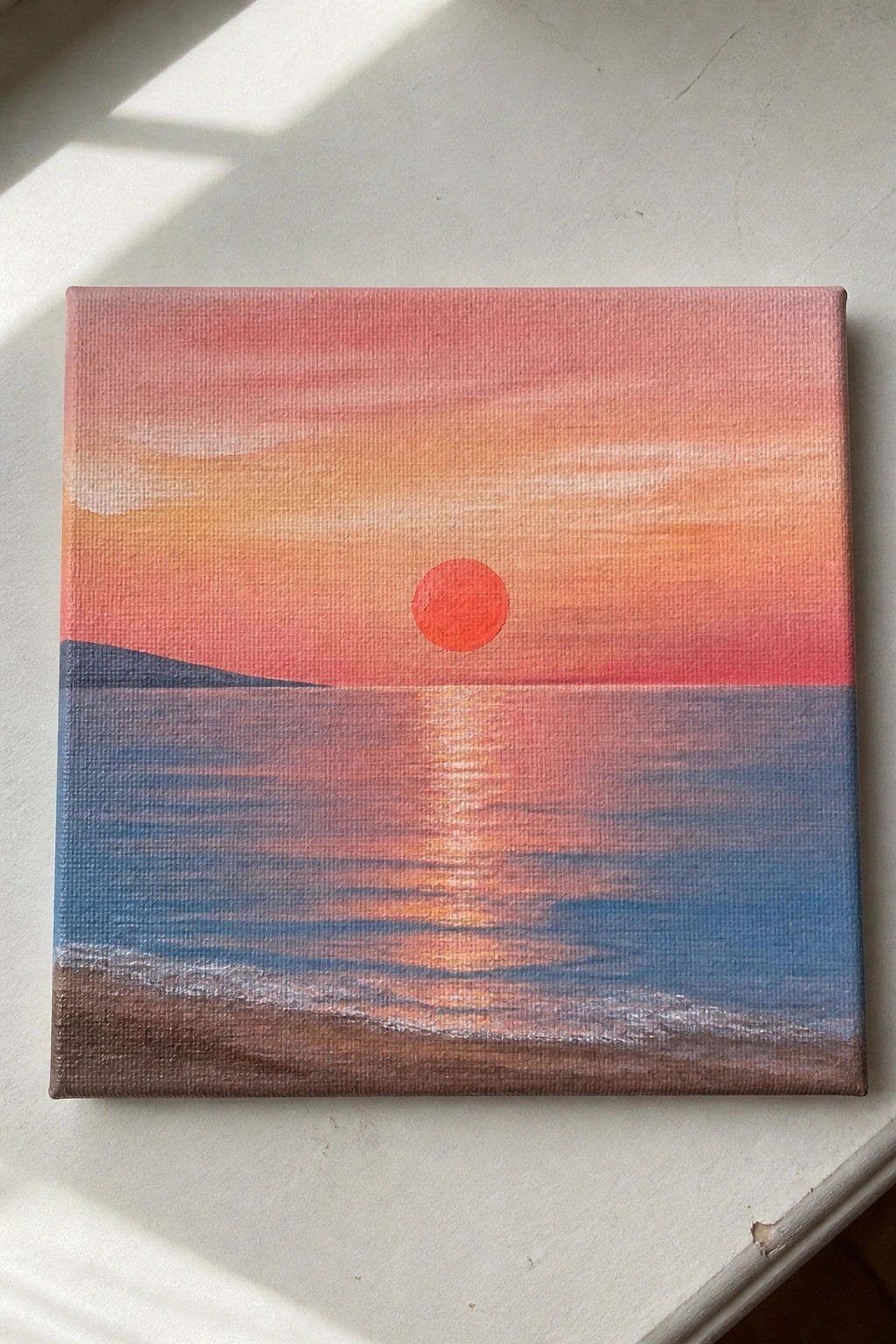 Red sun setting over ocean with pink-orange sky, water reflection, and shore