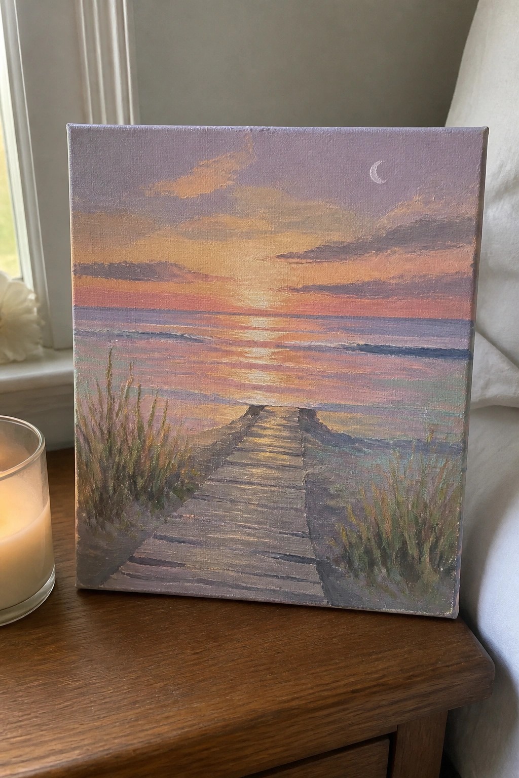 Sunset ocean painting with boardwalk, grasses, and crescent moon beside lit candle on wood