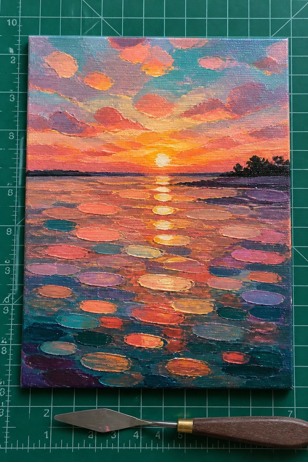 Vibrant sunset painting with colorful clouds, sun reflection, and lily pads beside palette knife