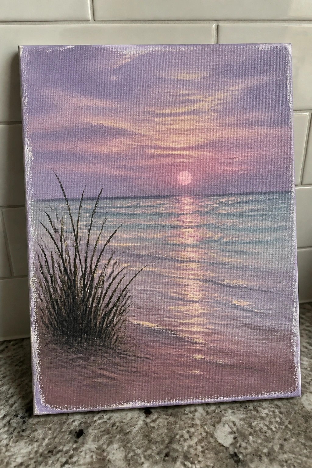 Acrylic painting of a sunset over the ocean with a pink sun, its reflection on the water, and dark grass on the left foreground beach.