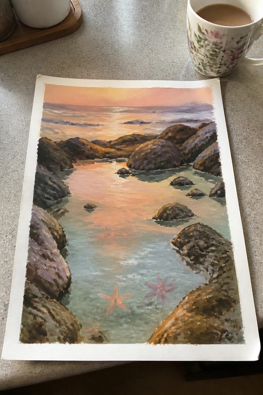 Watercolor sunset painting of rocky cove with starfish beside floral mug on countertop.