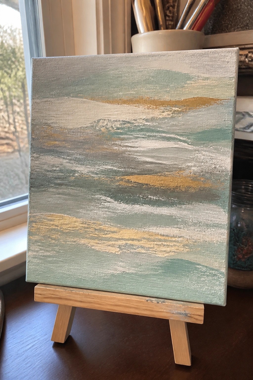 Abstract painting with green, teal, gray, white and gold brushstrokes on wooden easel
